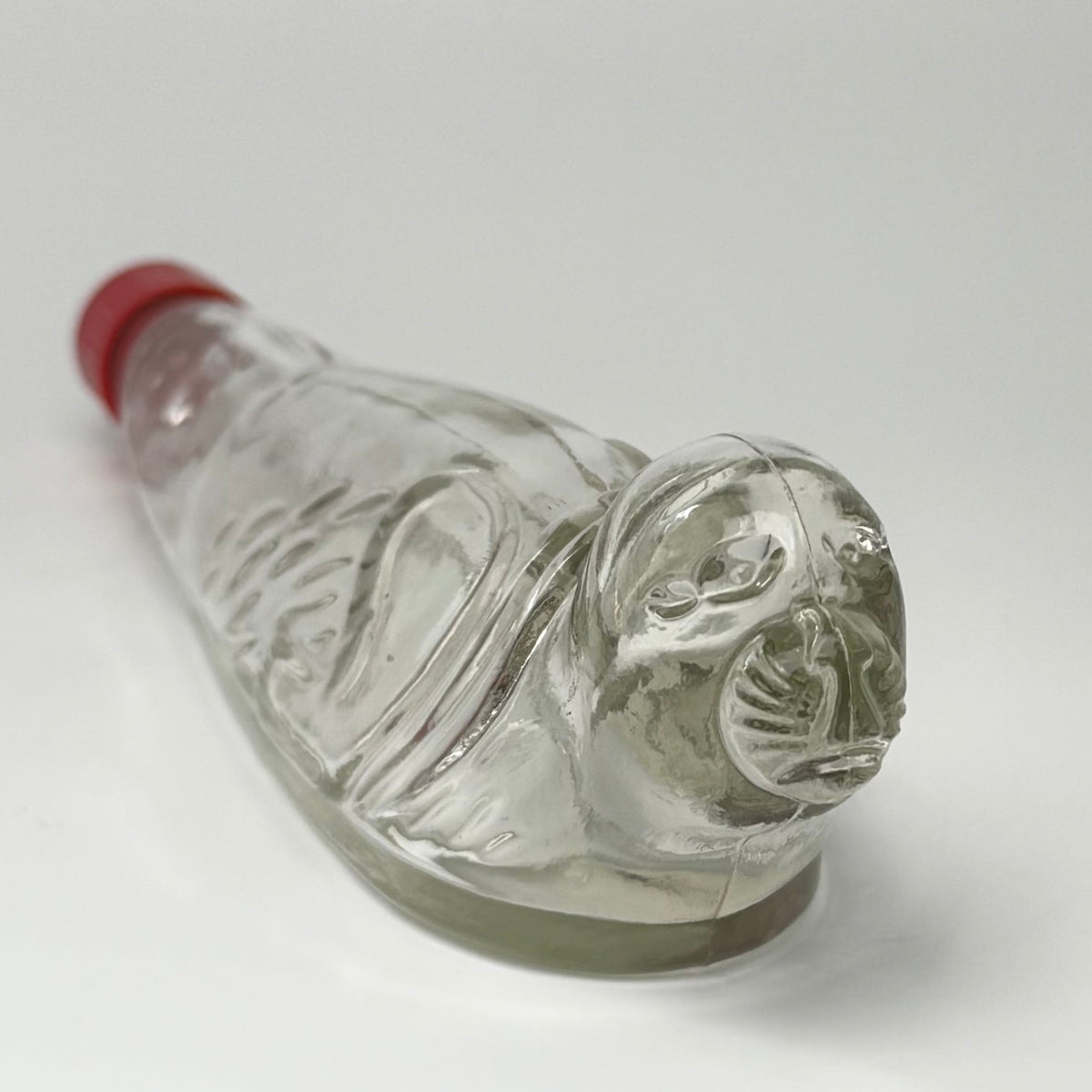 Vintage tysk figurflaske i klart formpresset glas, udformet som en liggende sæl – Seehund.

Flasken har detaljeret relief med sælens hoved, luffer og krop. Hoved og snude er udført i massivt glas.

Denne type figurflaske blev fremstillet i Tyskland som emballage til spiritus. Tilsvarende eksemplarer ses med originale etiketter mærket bl.a. “Seehund” og “Deutsches Erzeugnis”. Producenten af dette eksemplar er ikke identificeret med sikkerhed.

Med original/lignende ældre rød skrueprop i plast med kork på ind