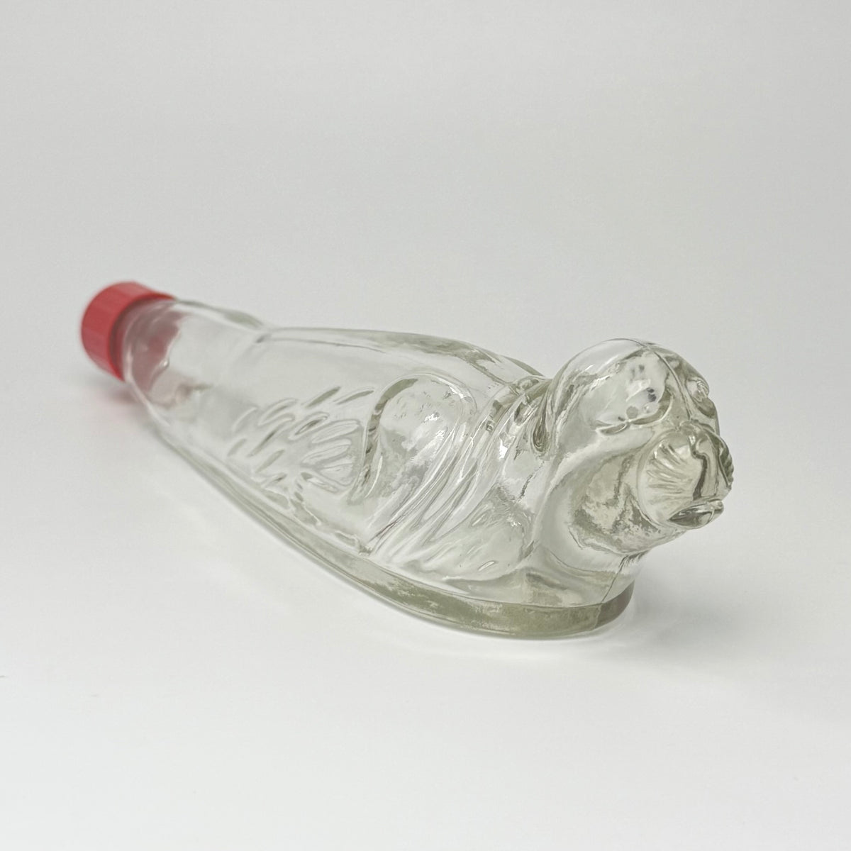 Vintage tysk figurflaske i klart formpresset glas, udformet som en liggende sæl – Seehund.

Flasken har detaljeret relief med sælens hoved, luffer og krop. Hoved og snude er udført i massivt glas.

Denne type figurflaske blev fremstillet i Tyskland som emballage til spiritus. Tilsvarende eksemplarer ses med originale etiketter mærket bl.a. “Seehund” og “Deutsches Erzeugnis”. Producenten af dette eksemplar er ikke identificeret med sikkerhed.

Med original/lignende ældre rød skrueprop i plast med kork på ind